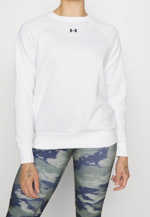 Osoba ubrana w białą, długorękawową bluzę Under Armour oraz niebiesko-zielone legginsy w wzory, stojąca na jednolitym tle.