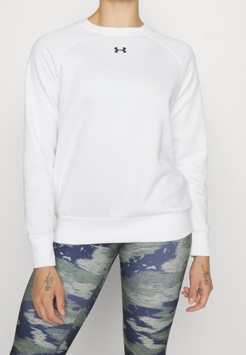 Personne portant un sweat-shirt blanc à manches longues Under Armour et un legging bleu-vert à motifs, debout devant un fond uni.