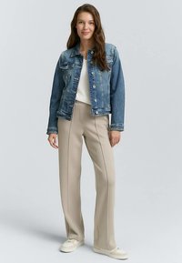 Chaqueta vaquera en azul medio, combinada con pantalones beige con costuras laterales. La modelo lleva una camiseta blanca y calzado claro, posando frente a un fondo neutro.