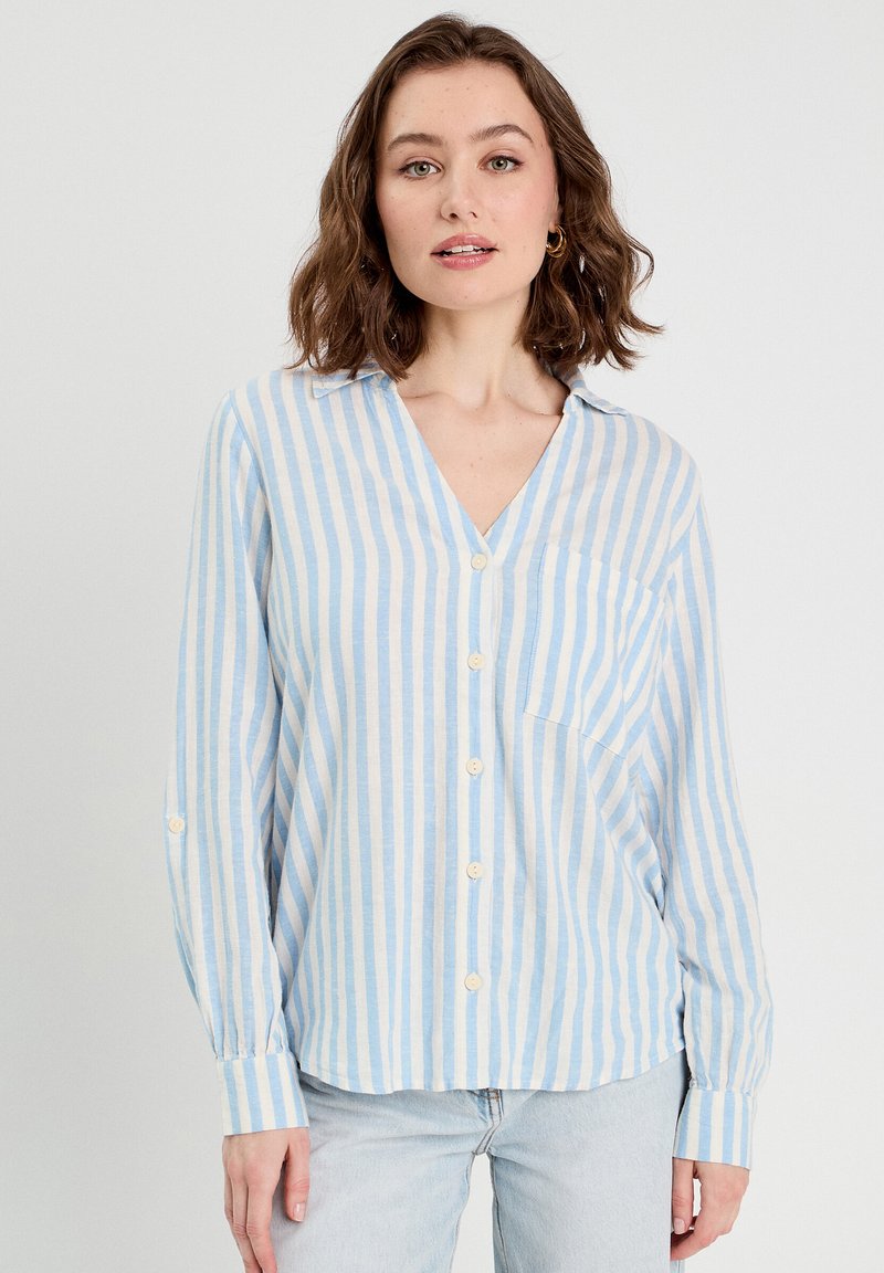 Chemise à rayures bleu clair et crème, avec un col en V, des manches longues, une poche poitrine et une coupe décontractée. Fabriquée en tissu doux.
