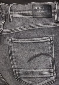Crne traperice imaju teksturiranu površinu, s velikim stražnjim džepom i kožnom oznakom s natpisom "G-Star RAW."