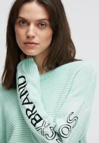 Hellgrüner, gestrickter Pullover mit gerippter Textur, versehen mit schwarzer Stickerei entlang des Ärmels. Lässige Passform mit weitem Ausschnitt.