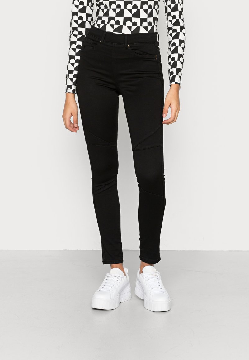 Calças pretas com um design justo e detalhes texturizados, apresentando costuras laterais e bolsos frontais, combinadas com ténis de plataforma brancos.