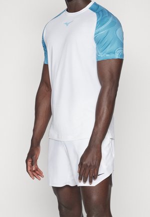 Mand iført hvid atletiktrøje med blå stribede ærmer og hvide løbeshorts, stående mod en ensfarvet baggrund.