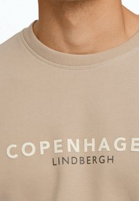 Felpa beige con collo rotondo e tessuto strutturato. Scritte bianche e scure che mostrano "COPENHAGEN" e "LINDBERGH." Finitura liscia.