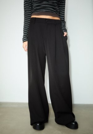 Person trägt hoch taillierte, weit geschnittene schwarze Hose, ein gestreiftes schwarz-graues langärmeliges Croptop und schwarze Plateauschuhe.