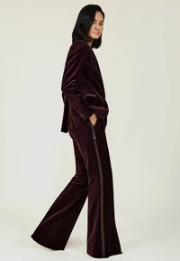 Burgundy bársonynadrág öltöny, szabott zakóval és bővülő nadrággal. A nadrág selyem oldalsávokkal rendelkezik, amelyek kontrasztot adnak, és sima tapintásúak.