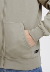 Sudadera con cremallera de color gris claro, con puños y dobladillo acanalados, grandes bolsillos frontales y una pequeña etiqueta de la marca en el lateral. Textura de tela suave.
