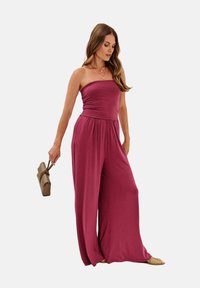 Jumpsuit bez ramínek v tmavě burgundské barvě, s přiléhavým korzetem a širokými kalhotami, vyrobený z měkké tkaniny. Doplněný béžovou kabelkou.