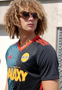 Camiseta de fútbol negra y verde azulado con acentos rojos, con el logotipo de "GALAXY". El modelo lleva gafas de sol y tiene el cabello rizado parcialmente visible.