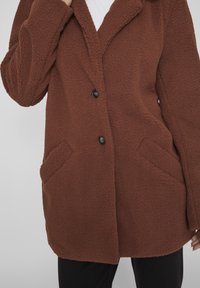 Brun, blød, tekstureret jakke med en reverskrave, to forlommer og to sorte knapper. Båret over en hvid top og sorte bukser.