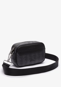 Borsa a tracolla nera compatta con motivo geometrico, logo Lacoste impresso e tracolla in tessuto regolabile su sfondo bianco.