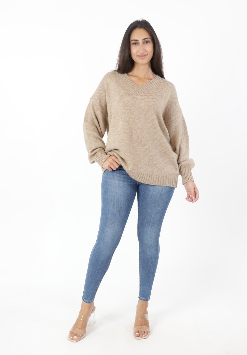 Beige oversized gebreide trui met een V-hals, gecombineerd met blauwe skinny jeans en transparante hakken. Basis, geweven stof met geribbelde manchetten.