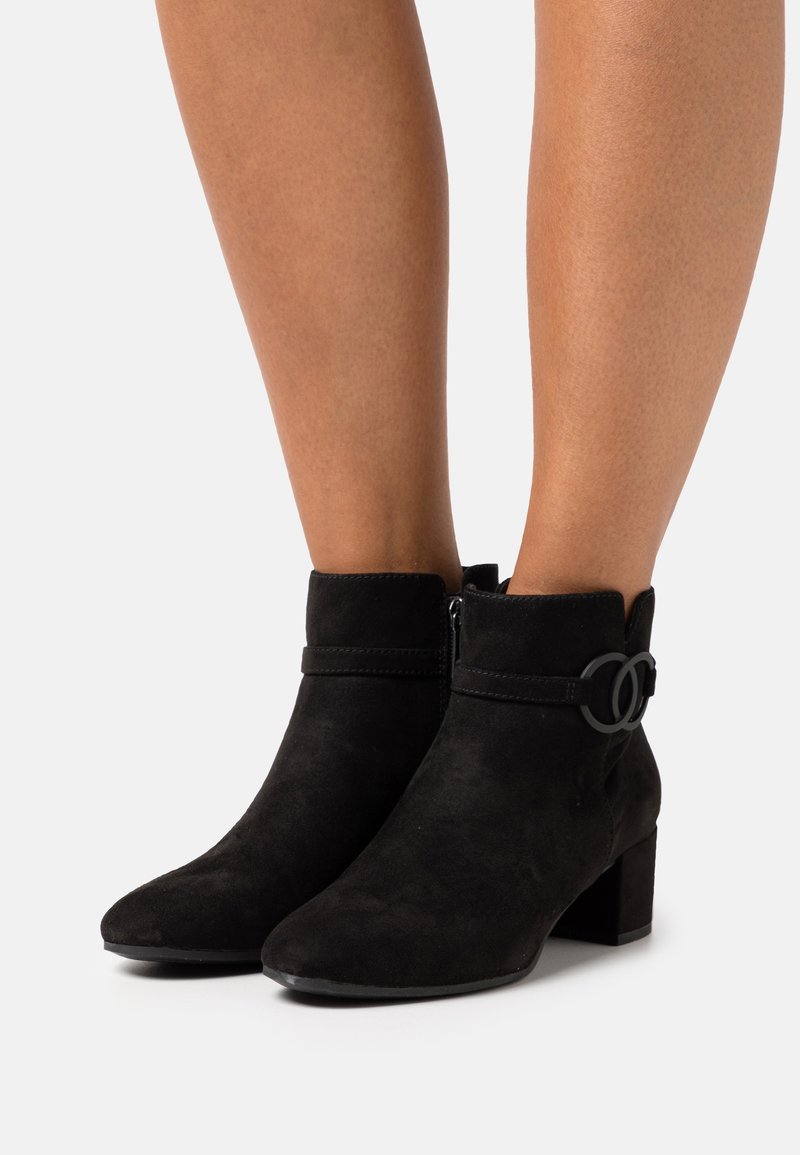 Bottines noires en daim avec talons carrés et boucle décorative latérale portées sur des jambes nues sur fond blanc.