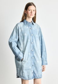 Heledest helesinises denimpuserdress, millel on nupud ees, pikad varrukad ja küljetaskud. Tootel on peen korduv logo muster üle kogu.