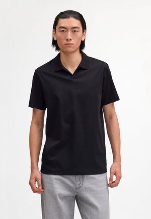 Mladi moški s ravnimi črnimi lasmi nosi kratko črno polo majico in svetlo sive kavbojke, stoji pred enobarvnim belim ozadjem.