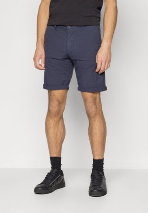 Granatowe bawełniane szorty z podwiniętymi mankietami, z zapięciem na guzik i paskiem, zestawione z czarnymi trampkami i ciemnymi skarpetkami.