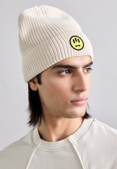 Berretto in maglia beige con patch a forma di faccina sorridente gialla, con una texture a costine e una vestibilità morbida. Design semplice, adatto per un look casual.