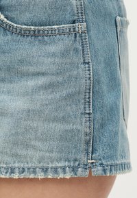 Φούστα από ανοιχτό μπλε τζιν με σχίσιμο μπροστά, εμφανείς ραφές και ακατέργαστο ξεφτισμένο τελείωμα, φορεμένη σε άτομο με φόντο απλό λευκό.