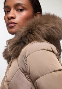 Giacca puffer beige con cappuccio in pelliccia marrone removibile. La texture trapuntata e la chiusura con zip accentuano il design. Finitura in tessuto liscia.