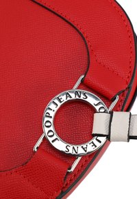 Borsa a forma di cuore in pelle rossa con superficie testurizzata, dotata di un accento in metallo circolare argentato e di una tracolla beige. Dettagli di bordatura nera visibili.