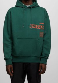 Sweat à capuche vert avec une poche avant. Graphique "NIRVANA" en orange sur la poitrine et la manche. Tissu texturé avec une coupe décontractée.