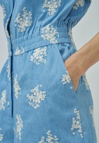 Mono de mezclilla azul con bordado floral blanco, cinturilla elástica y bolsillos. Cuenta con un frente abotonado y una textura suave.