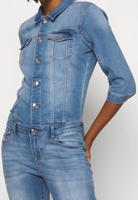 Džínový overal s přiléhavým střihem, světle modré barvy, tříčtvrtečním rukávem a kovovými knoflíky, který má přední kapsy a límec.