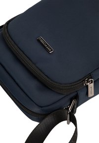 Borsa in tessuto blu navy con una superficie liscia, dotata di chiusura con zip e manico nero. Etichetta logo metallica sul davanti. Design compatto.