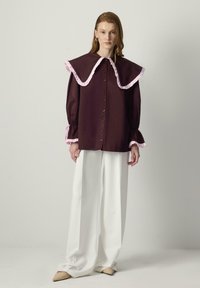 Bordeauxkleurige blouse met knopen en een grote rozenkrans met plooien aan de kraag en manchet, gecombineerd met witte brede benen broek en beige puntige schoenen.