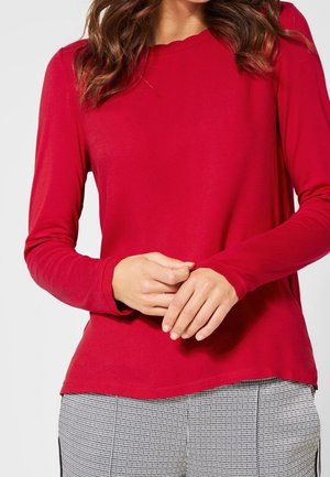 Woman wearing a red long-sleeve shirt and black-and-white checkered pants, hands clasped in front.