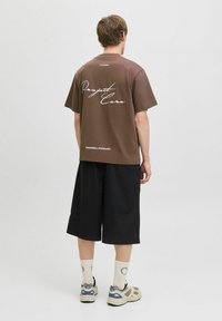 Brun bomulds t-shirt med løs pasform, med hvid tekst "Project Core" på ryggen, parret med sorte shorts og beige sneakers.
