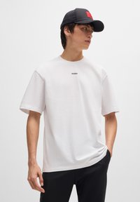 Vitt bomull T-shirt med korta ärmar, med en svart "HUGO" logotyp på bröstet. Bärs med svarta byxor och en svart keps.