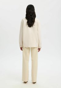 Femme aux longs cheveux foncés portant une chemise à manches longues ivoire et un pantalon à jambes larges, debout de dos sur un fond blanc uni.