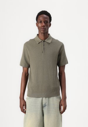 Olive green short-sleeve polo shirt with a textured knit design, three-button placket, and ribbed hem, paired with light denim shorts.