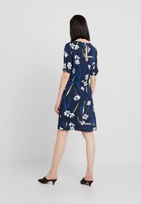 Laivansinis kukkamekko lyhyillä hihoilla, jossa on valkoisia kukkia ja vihreitä yksityiskohtia, vyötäröside ja takana avonainen yksityiskohta. Sileä kangas.