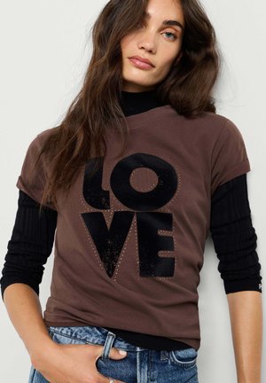 Frau mit langen braunen Haaren trägt ein braunes T-Shirt mit schwarzen "LOVE" Buchstaben über einem schwarzen Langarmshirt und blaue Jeans.