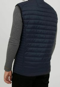 Navy blue padded vest with horizontal stitching, lightweight fabric, high collar, and fitted design. Worn over a grey long-sleeve shirt.
