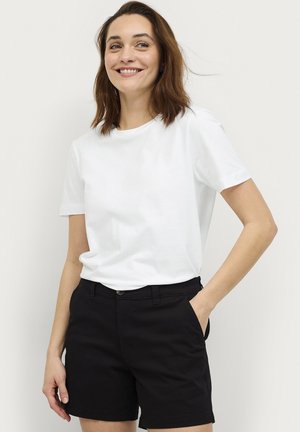 Ung kvinde med skulderlangt brunt hår iført en hvid t-shirt med korte ærmer og sorte shorts, smilende med den ene hånd i lommen.