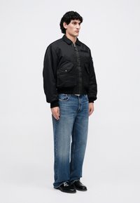 Sort bomberjakke med lynlås foran, to forreste lommer og ribbede manchetter, parret med slidte blå lige bukser og sorte sko.