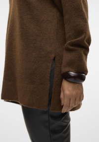 Brown knit sweater with side slits, soft texture, and loose fit. Hand with brown bangle rests against the side, paired with black pants.