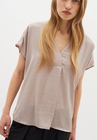 Blusa beige chiaro con scollatura a V, maniche corte e vestibilità ampia. Il tessuto presenta una finitura morbida e leggermente testurizzata.