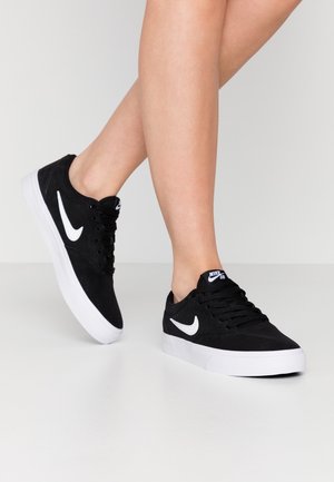 Nogi w czarnych butach Nike SB z białym logo i podeszwami, stojące na jednolitej, jasnej powierzchni i tle.