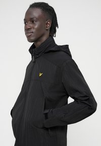 Black hooded jacket with a textured fabric, featuring a zip closure, side pockets, and a small yellow bird logo on the chest.