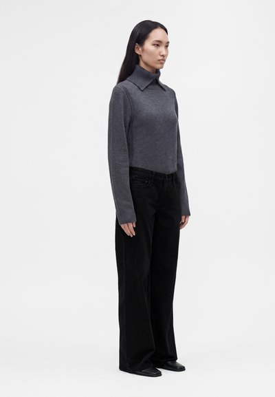Femme aux longs cheveux noirs, debout, portant un pull à col roulé gris et un pantalon noir ample sur un fond clair uni.