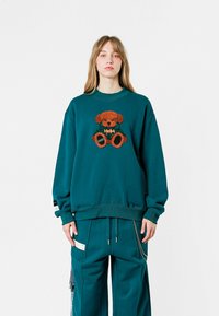 Sudadera de gran tamaño en color teal con un diseño de osito de peluche marrón y esponjoso en la parte frontal, con puños y dobladillo acanalados, combinada con pantalones teal a juego.