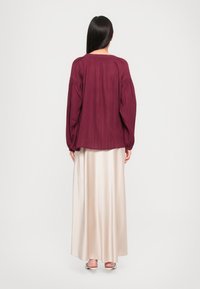 Femme portant une longue jupe beige, un blouse à manches longues bordeaux et des sandales à talons blancs, se tenant de dos sur un fond uni.