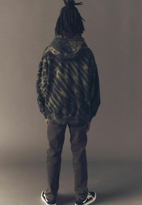 Kapuzensweatshirt im Batikdesign in dunkelgrün und schwarz, kombiniert mit dunklen Hosen und schwarz-weißen Sneakers. Ansicht von hinten.