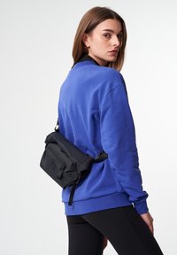 Schwarze Umhängetasche, die über einem blauen Sweatshirt getragen wird, mit schlichtem Design, Reißverschluss und strukturiertem Stoff. Befestigter schwarzer Riemen.