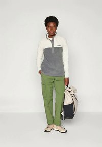 Πουλόβερ fleece σε κρεμ και γκρι, με γιακά που κουμπώνει; πράσινες cargo παντελόνες; κρατώντας ένα ανοιχτόχρωμο, διπλωμένο σακίδιο; casual παπούτσια.